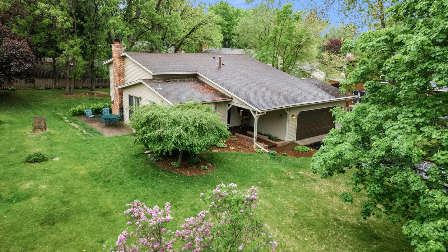 Aerial view of a beige single-family home with a brown roof, attached garage, brick chimney, and green lawn with trees.