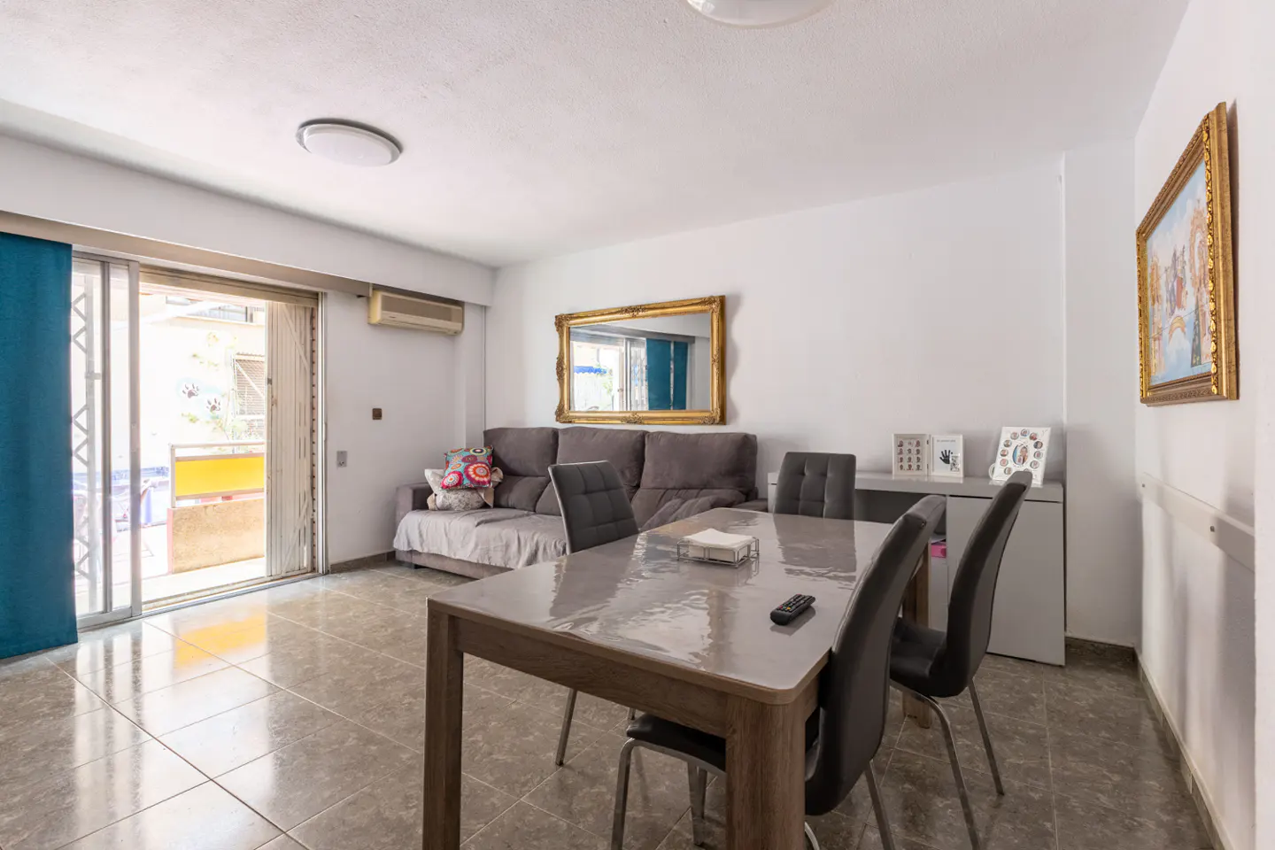 Living room with a dining table, gray sofa, and balcony. The walls are white, and the floor is tiled. A gold-framed mirror hangs above the sofa.