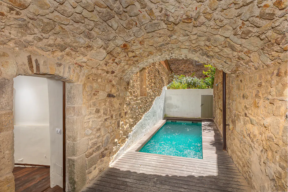 Indoor pool with stone walls and arched ceiling. Wooden deck surrounds the turquoise water. Doorway to interior room with wood floors.