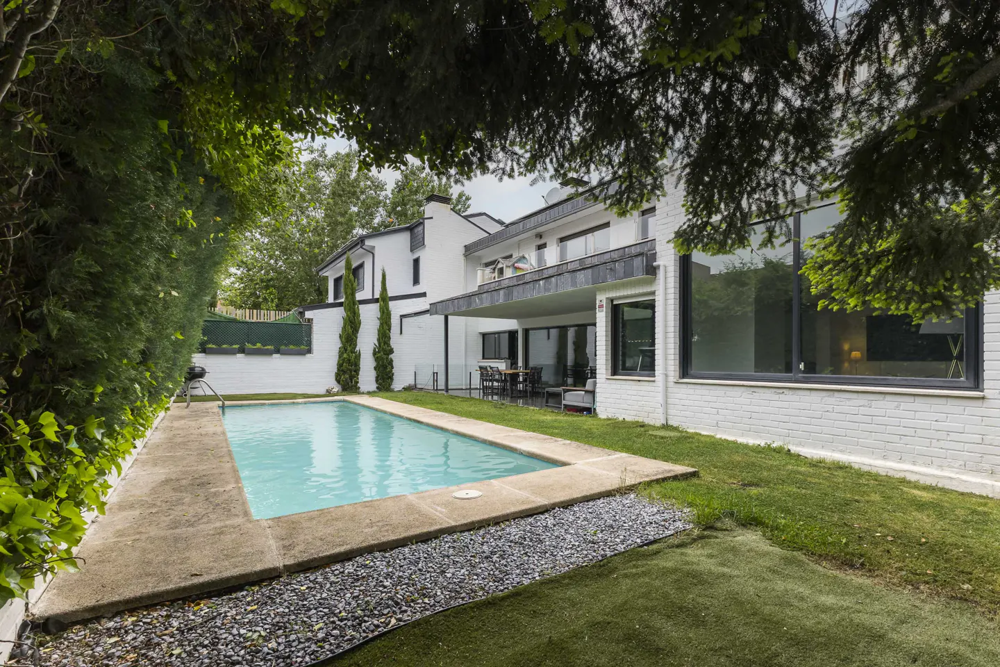 A backyard with a rectangular pool, stone patio, and white brick house with large windows.