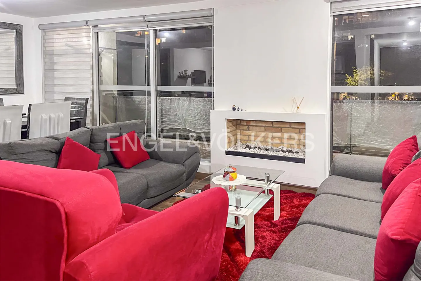 Modern living room with gray sofas, bright red cushions, a glass coffee table, and a fireplace on a red shag rug.