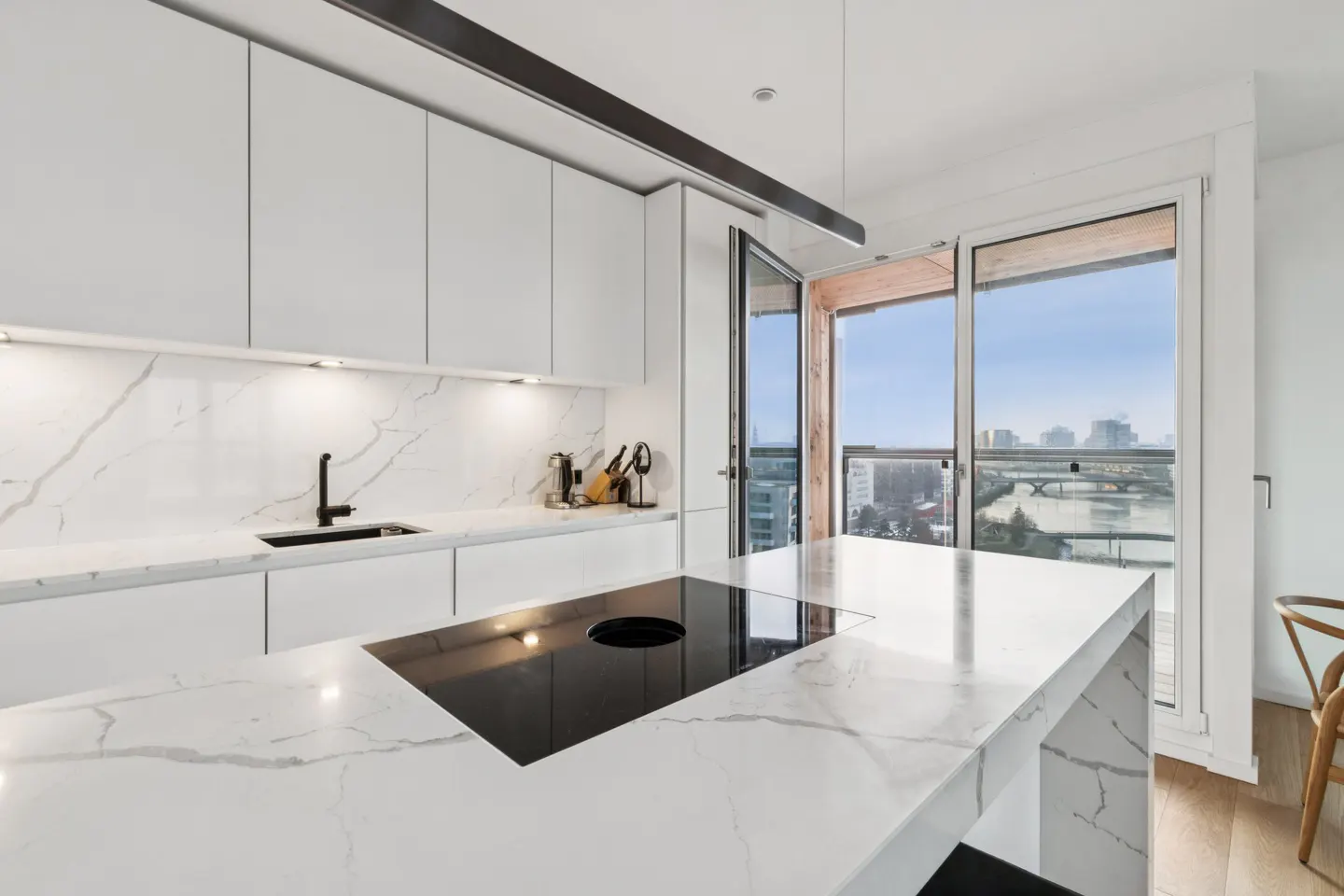 Bright, modern kitchen with white cabinets, marble countertops, and a black cooktop island. Large windows offer a city view.