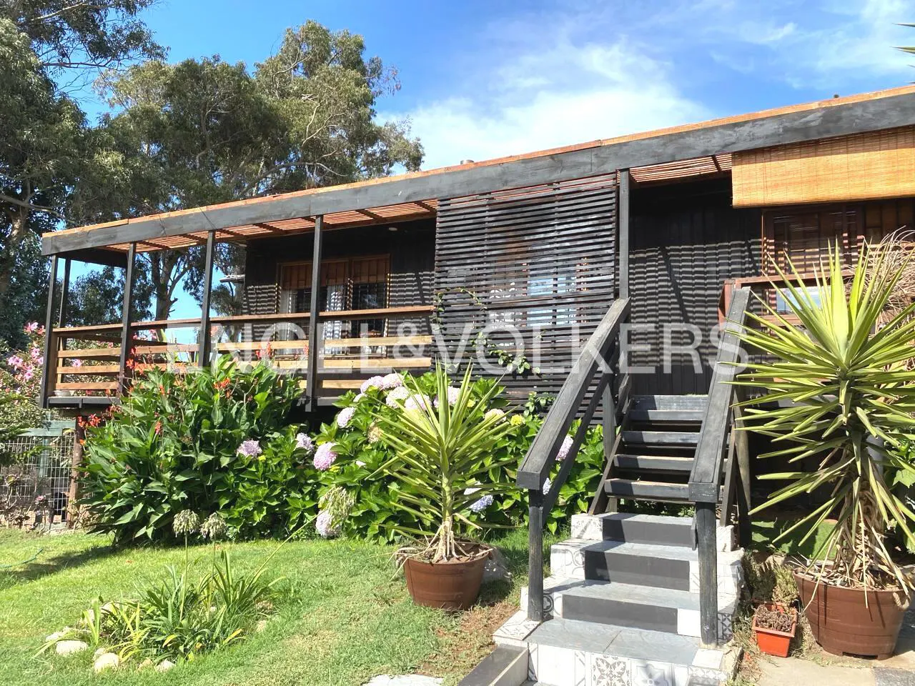 Exterior of a dark wood house with a porch, stairs, and lush green plants. Blue sky in the background.