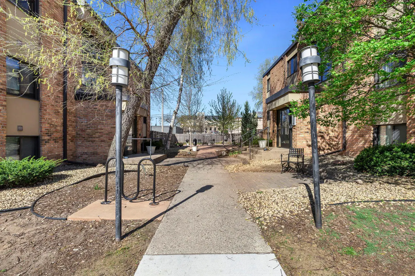 Exterior view of a brick apartment complex with a walkway, trees, and lamp posts on a sunny day.