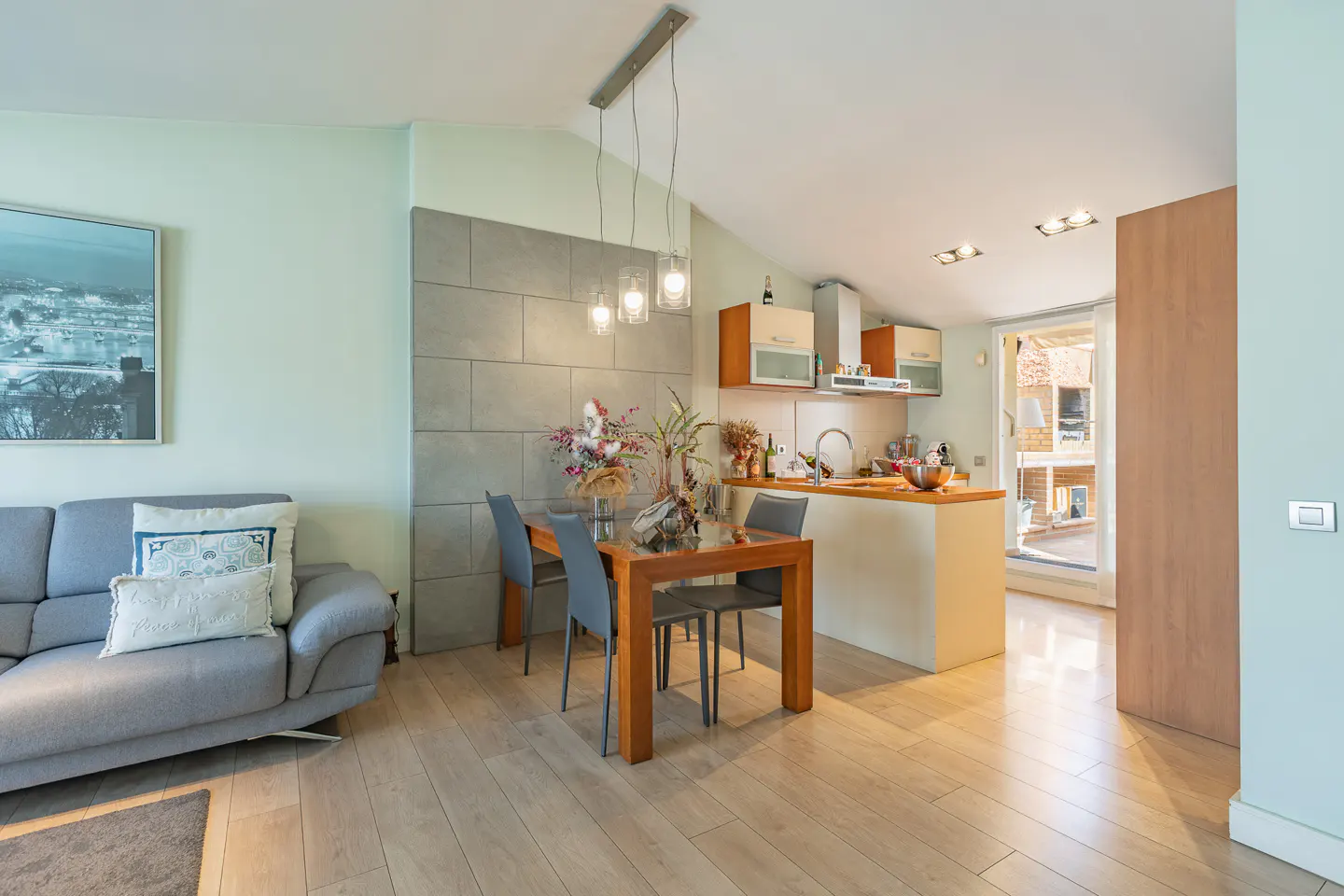 Open-concept living space with a gray sofa, wood dining table, and kitchen island. Light wood floors and pale green walls create a bright, airy feel.