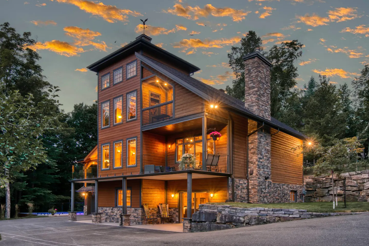 Three-story brown house with stone chimney and balconies, lit by warm lights against a sunset sky.
