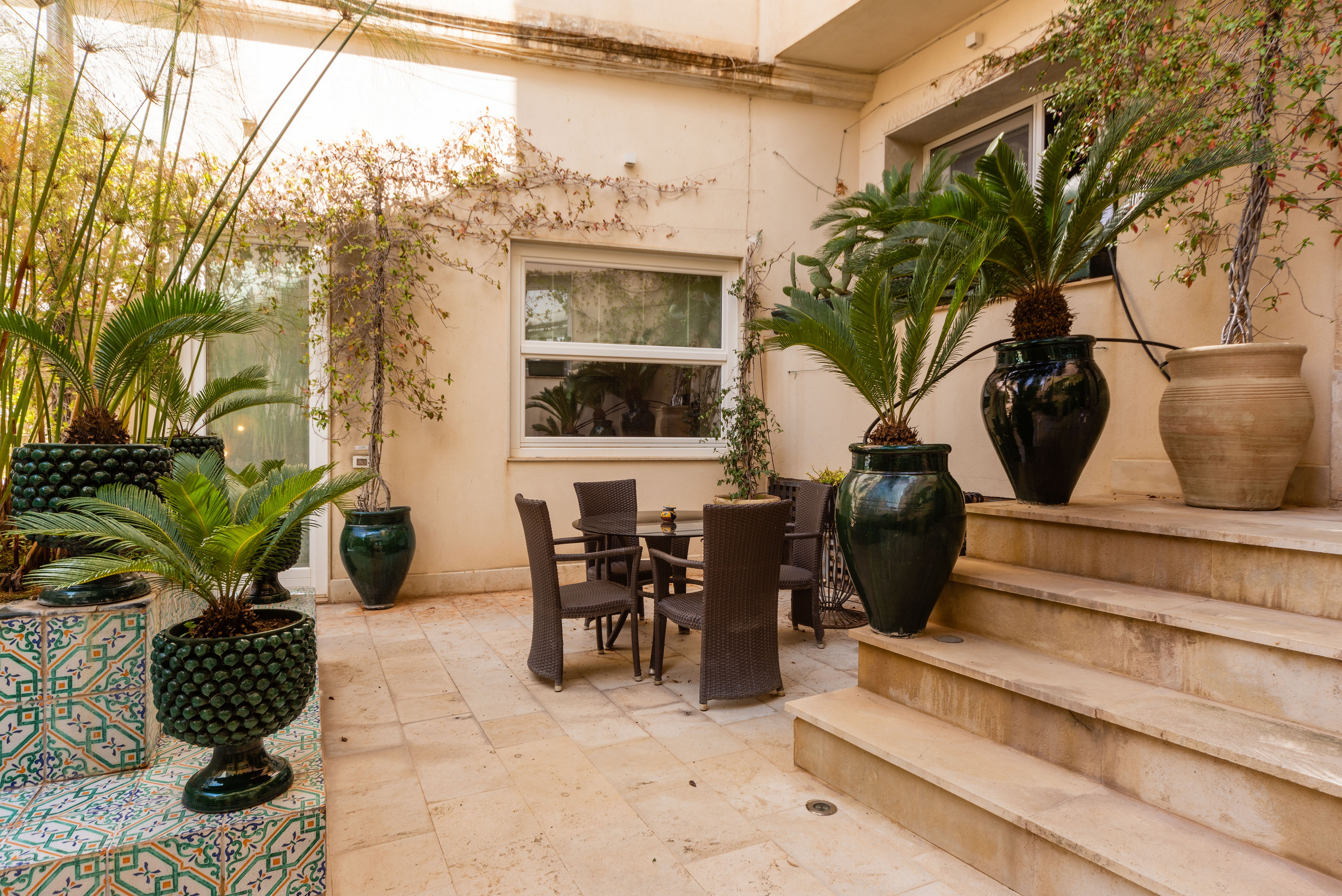 Outdoor patio with a table and chairs, potted plants, and stone steps. The walls are beige with climbing vines.