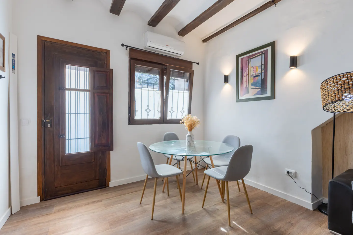 A dining area with a glass table, four gray chairs, and a rustic wooden door. Beamed ceiling and wood floors add warmth.