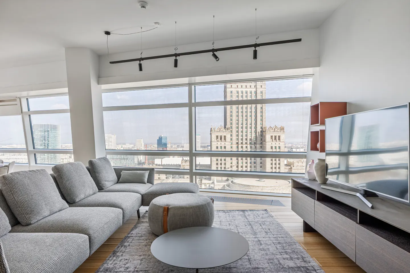 Bright living room with a gray sectional sofa, round coffee table, and a large window with a city view.