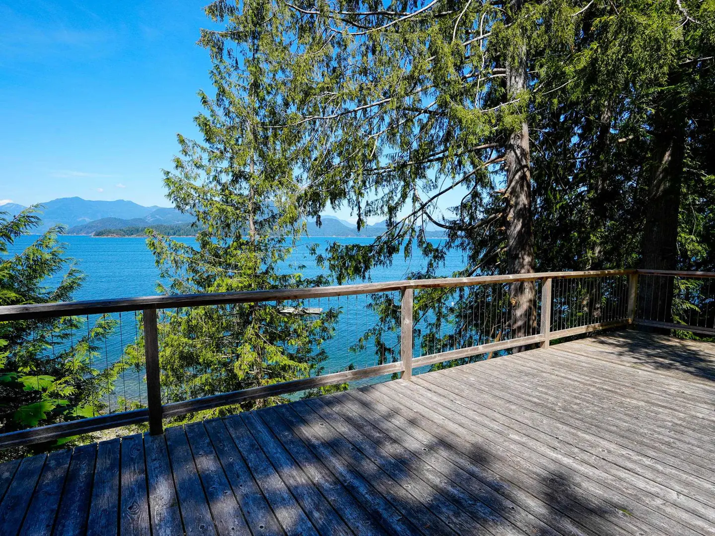 Wooden deck with railing overlooks blue ocean and islands on a sunny day. Trees frame the view.