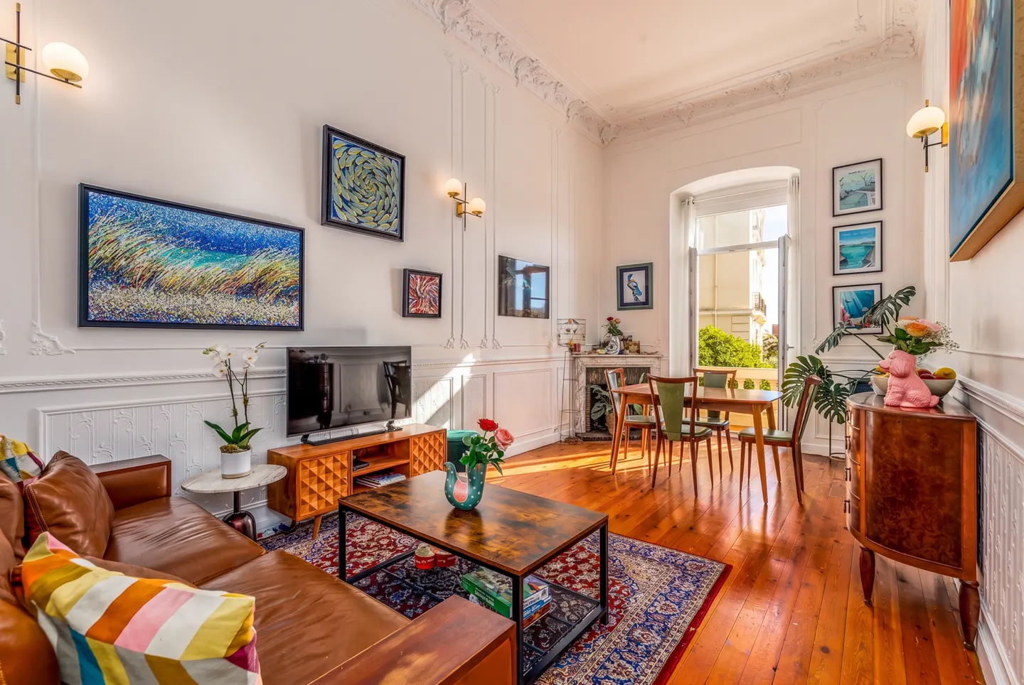 Bright living room with leather sofa, wooden coffee table, dining area, and ornate white walls with colorful art.