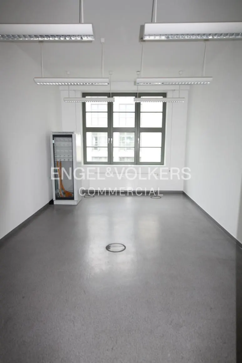 Empty office space with gray floors, white walls, and fluorescent lighting. A window and server rack are visible.