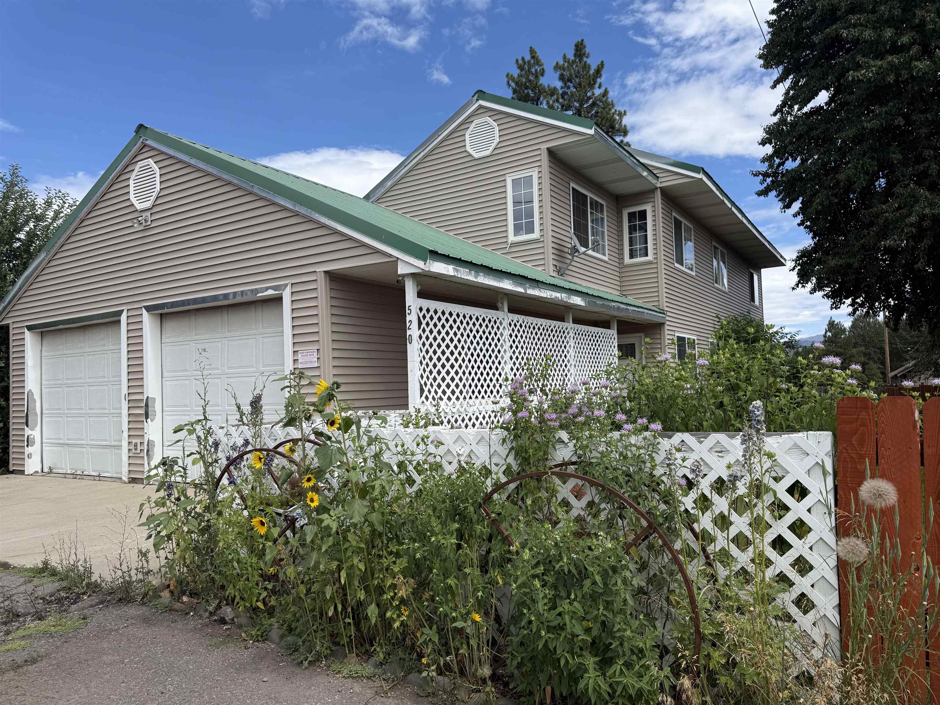 Two-story beige house with a green roof, two-car garage, and a white lattice fence surrounded by sunflowers and greenery.