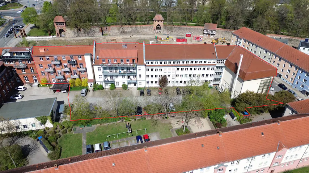 Aerial view of a multi-story apartment complex with red roofs, surrounded by green spaces, parking, and a historic stone wall in the background.