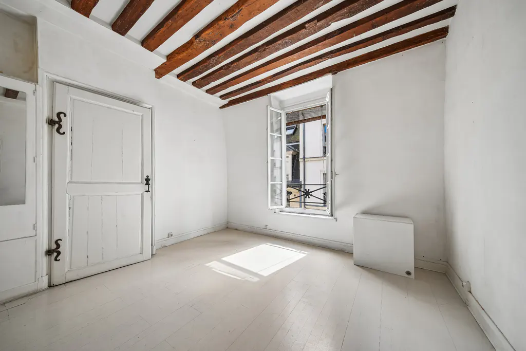 Bright, empty room with white walls, wood floors, and exposed ceiling beams. A window is open, and a white door is on the left.