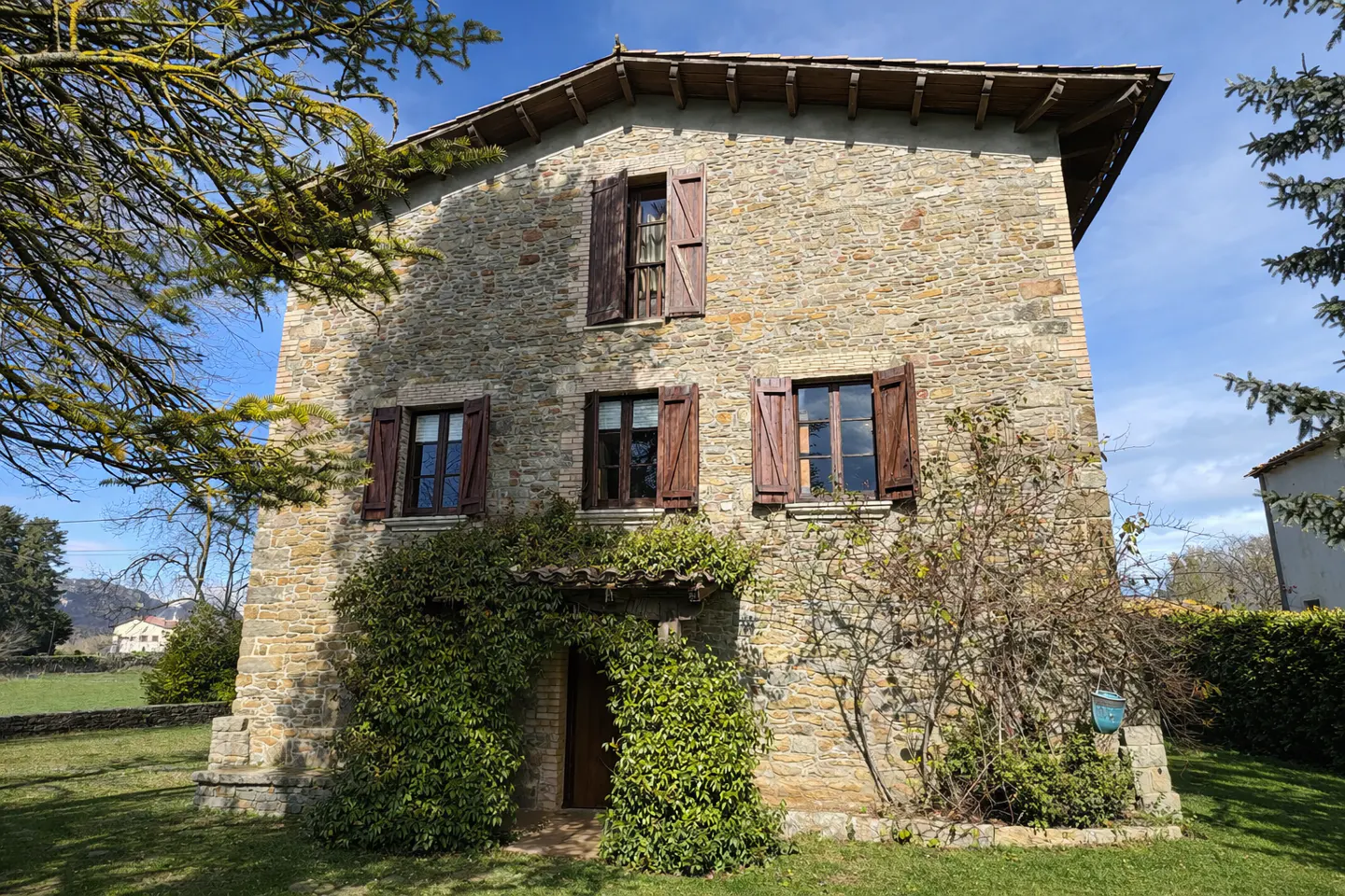 Two-story stone house with brown shutters and a green vine-covered entrance on a sunny day.