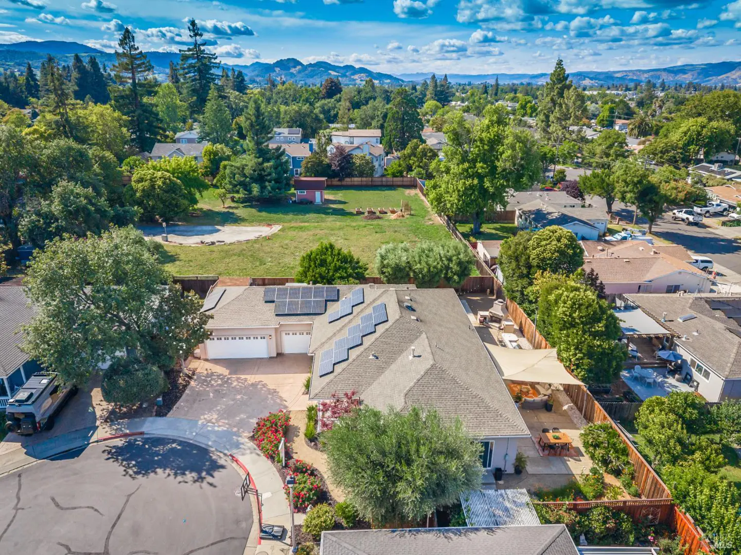 Aerial view of a suburban home with solar panels, a large backyard, and a patio with a shade sail under a blue sky.