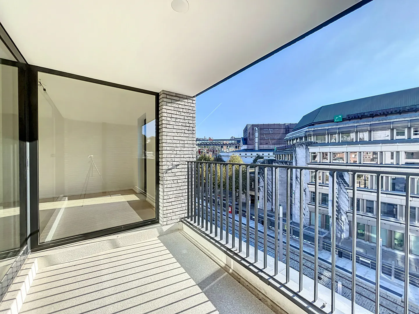 Balcony view with black metal railing, brick wall, and large glass door. City buildings are visible in the background under a blue sky.