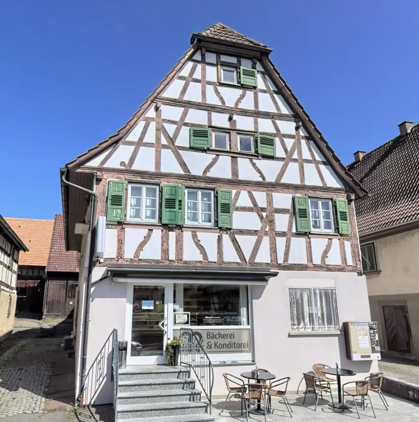 Two-story bakery with a half-timbered facade, green shutters, and outdoor seating on a sunny day.