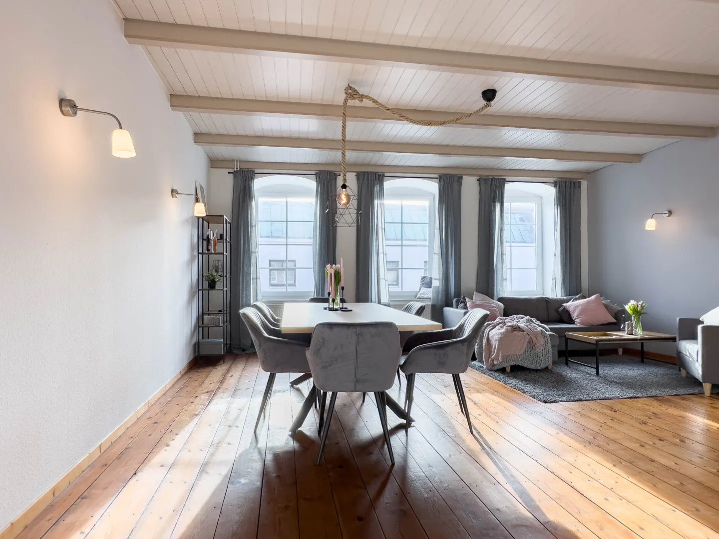 Bright, open-concept living and dining area with wood floors, white ceiling beams, and gray furniture. A rope light fixture hangs above the dining table.
