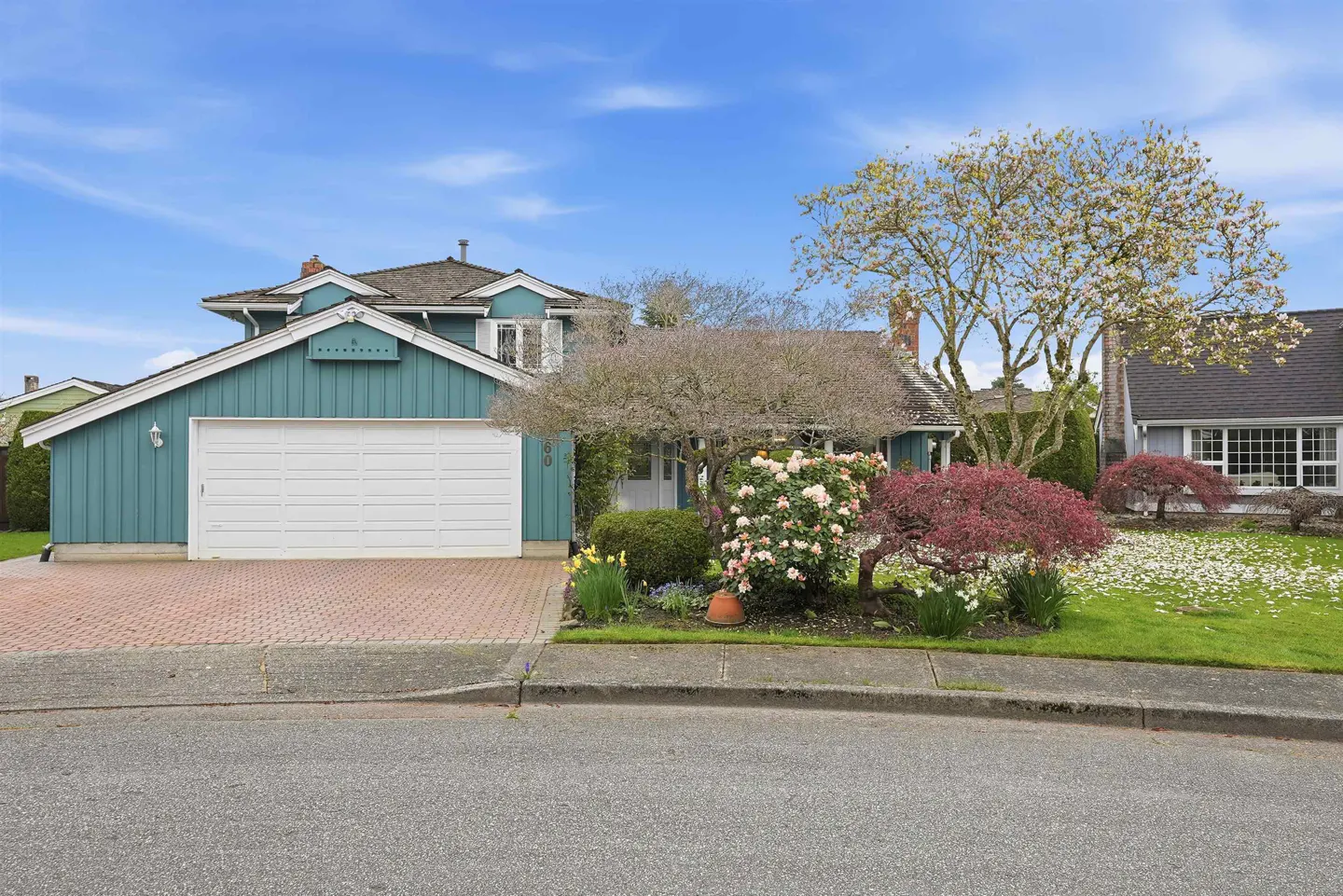 Exterior of a two-story teal house with a white garage door, brick driveway, and blooming trees.