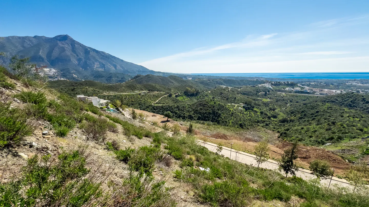 Scenic view of green hills, a mountain, and the ocean under a blue sky. A road winds through the landscape.
