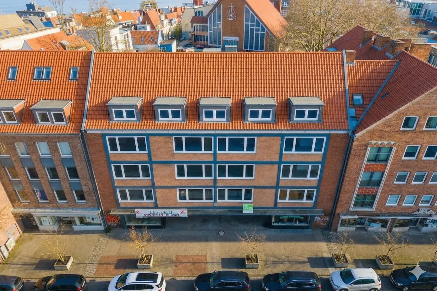 Aerial view of a three-story brick building with a red tile roof and gray dormers. Cars are parked on the street below.