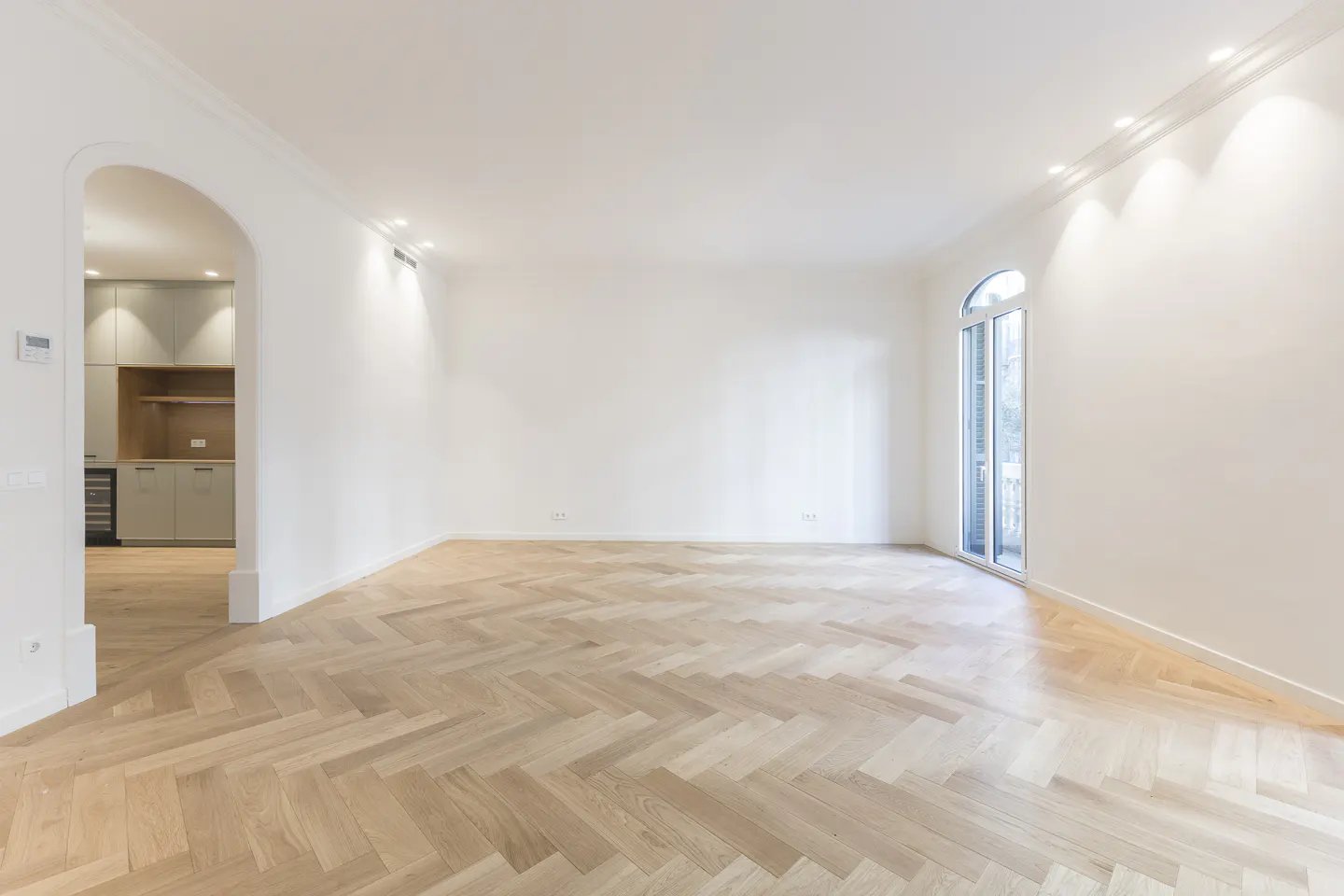 Bright, empty room with herringbone wood floors, white walls, and arched doorway to kitchen. Arched window lets in natural light.
