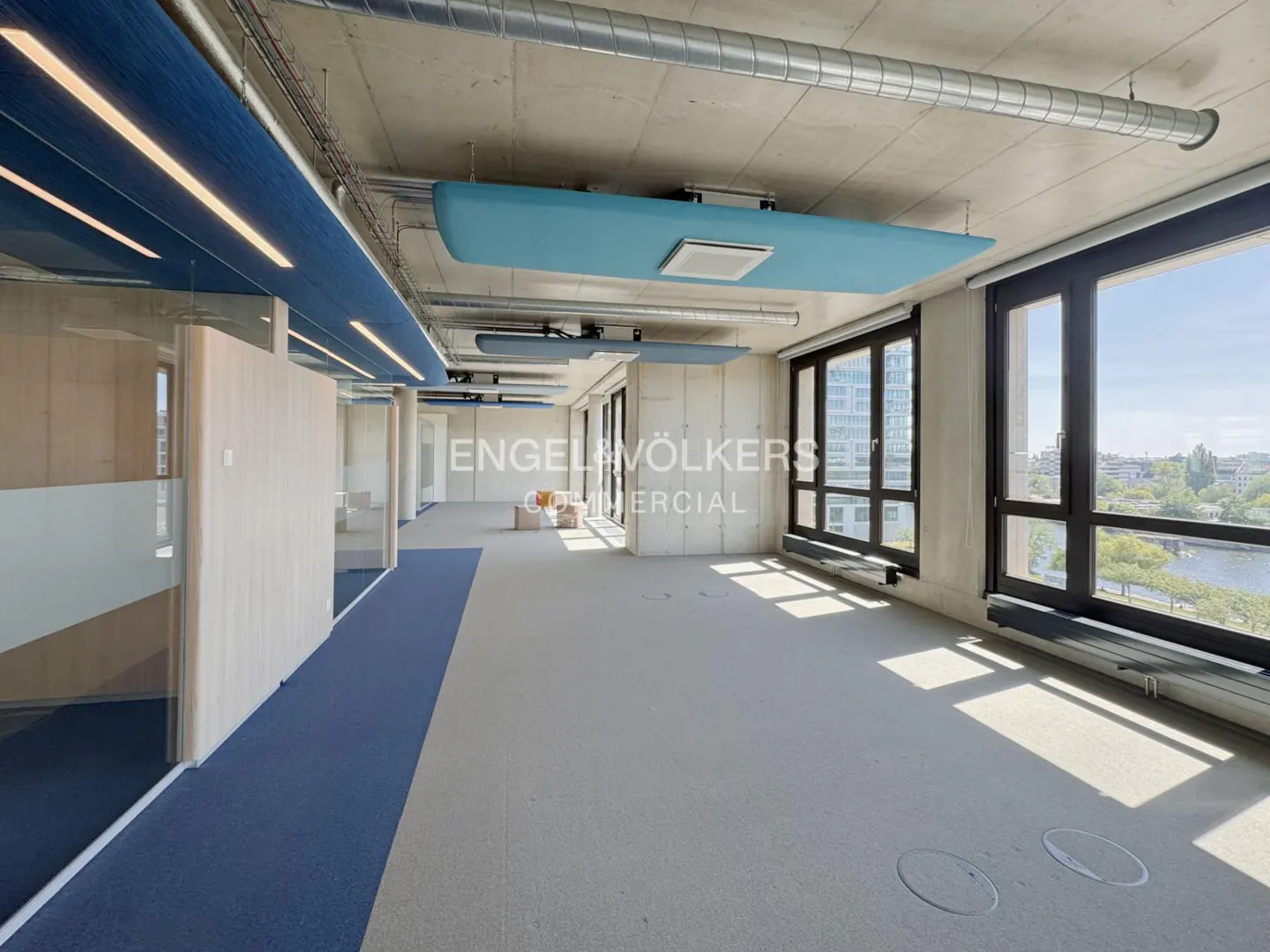 An empty office space with blue ceiling panels, large windows, and a view of a city and river.