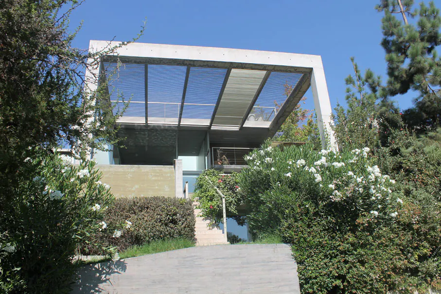 Modern home with a concrete frame and slatted roof, surrounded by lush greenery and a clear blue sky.