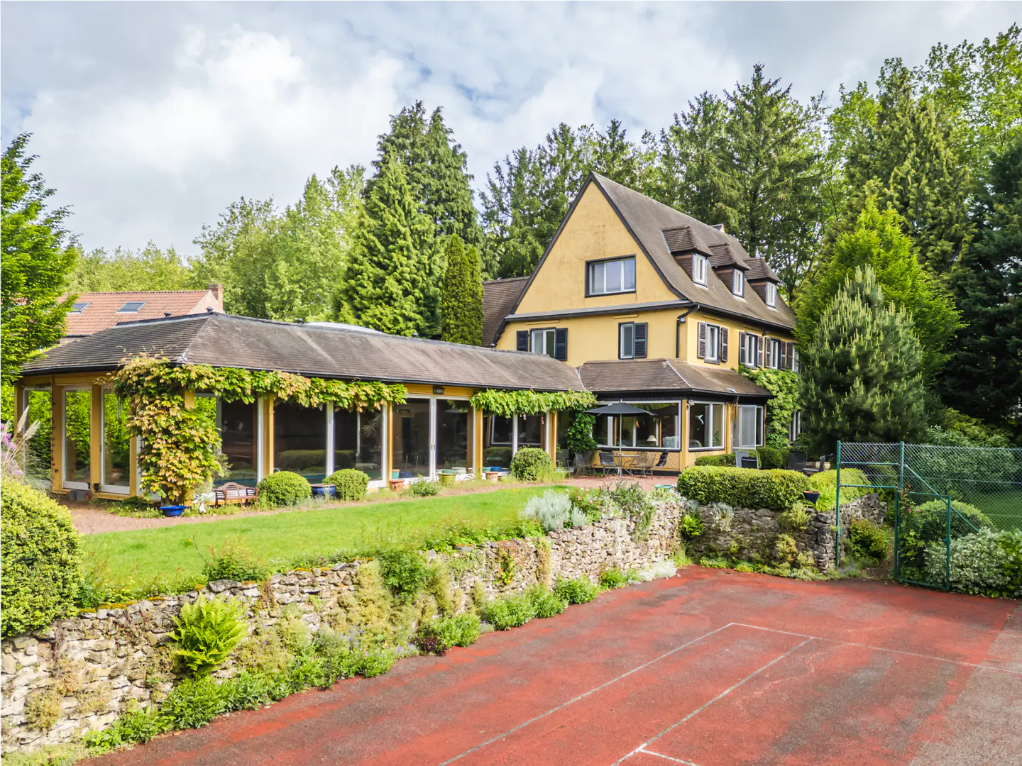 Exterior view of a yellow two-story house with a red tennis court and a glass-enclosed porch. Tall green trees surround the property.