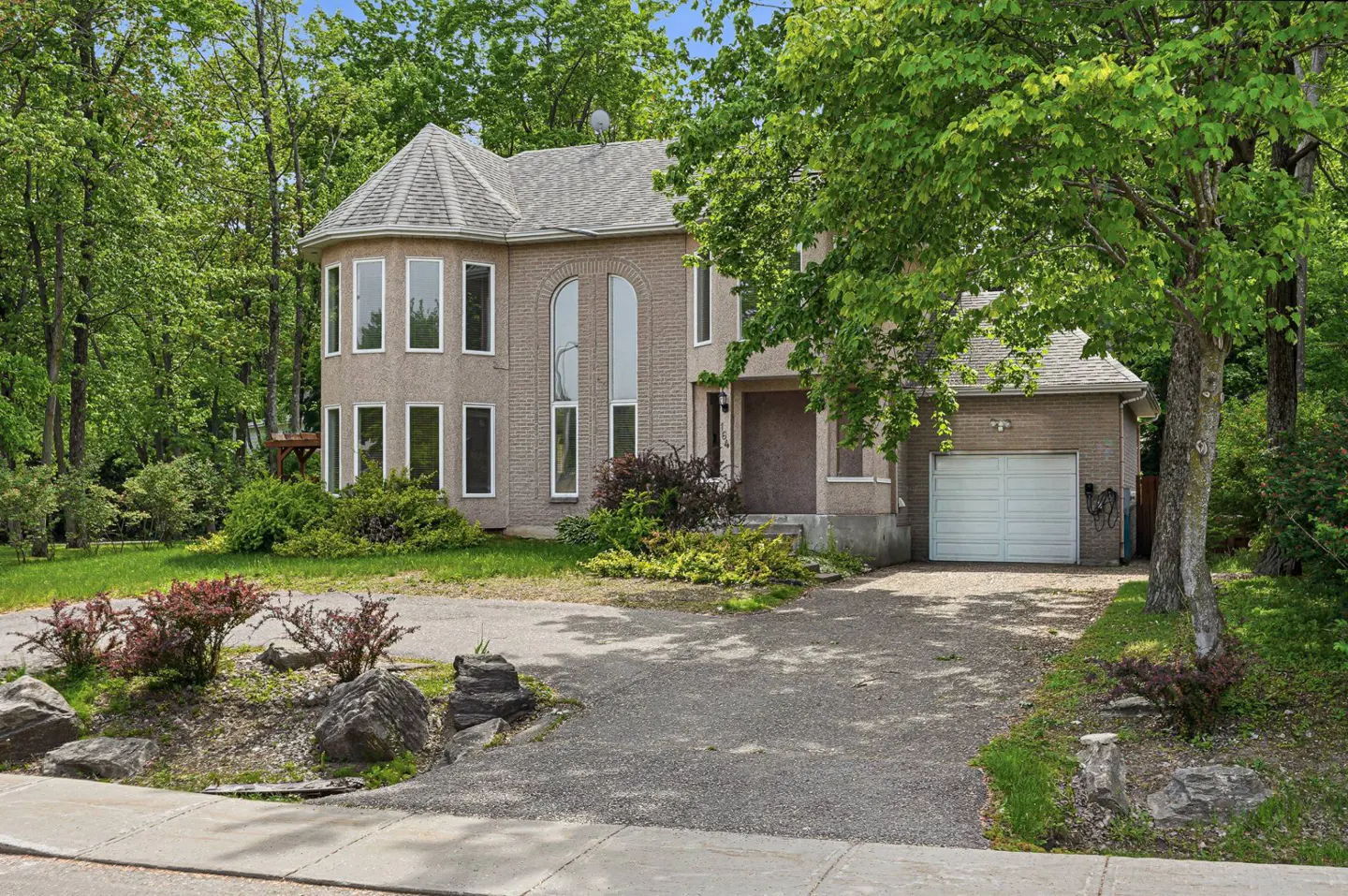 Two-story tan brick house with a turret, arched windows, and a white garage, surrounded by lush trees and a gravel driveway.
