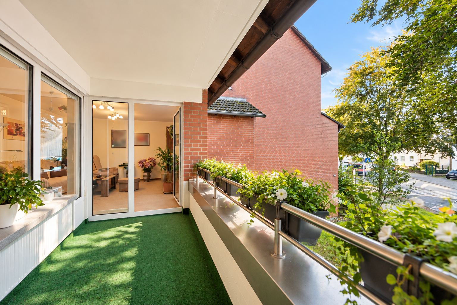 Balcony with green turf flooring, flower boxes on a metal railing, and an open glass door leading into a bright living room. [AI-generated text]