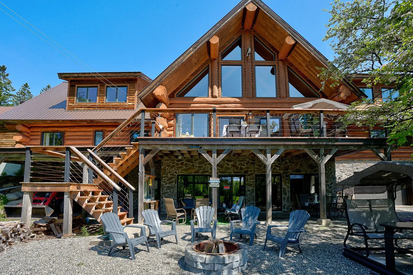 Exterior view of a two-story log cabin with a stone foundation and a gravel patio with a fire pit.