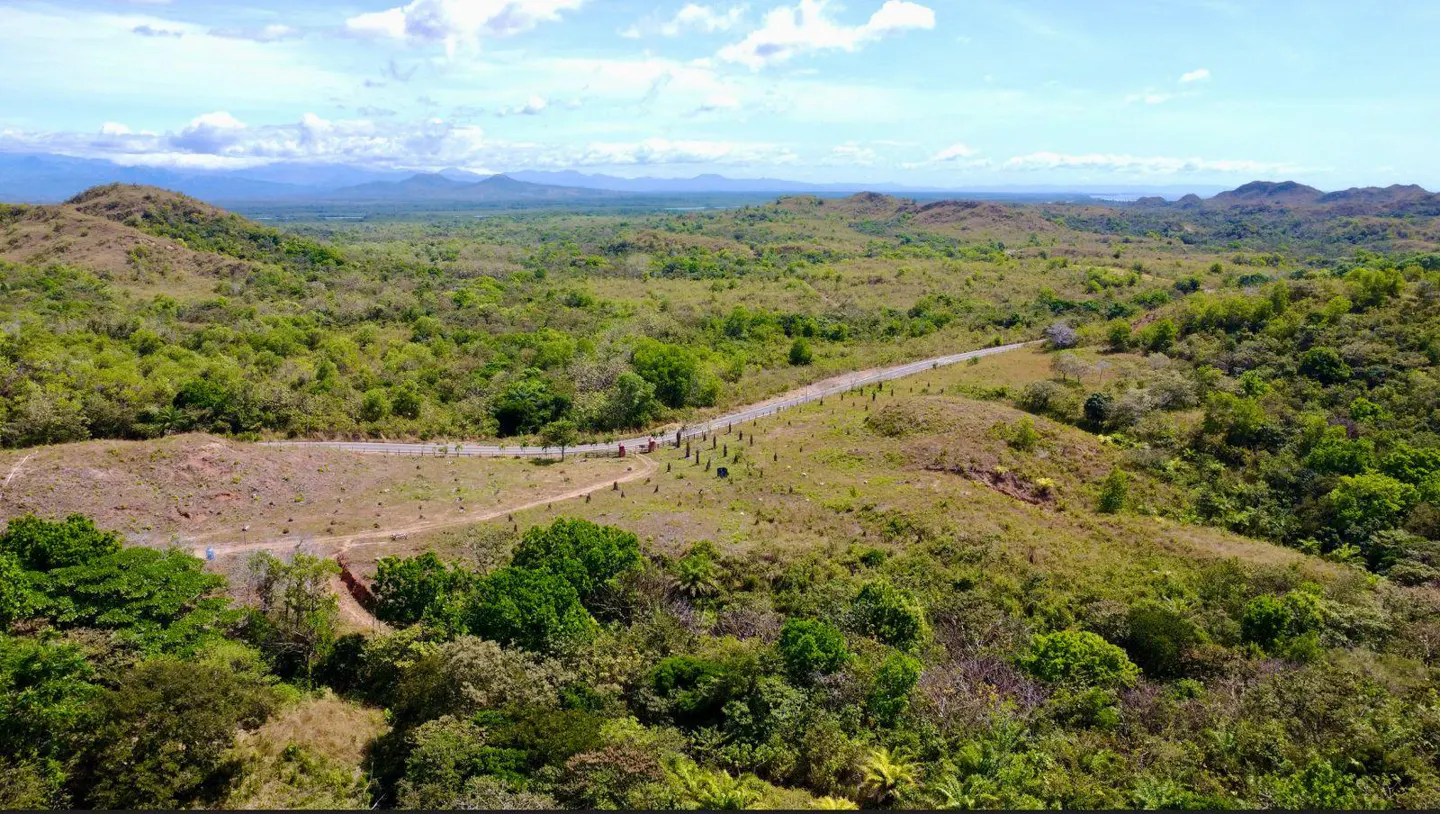 Aerial view of a green, hilly landscape with a road winding through it under a blue sky with white clouds.