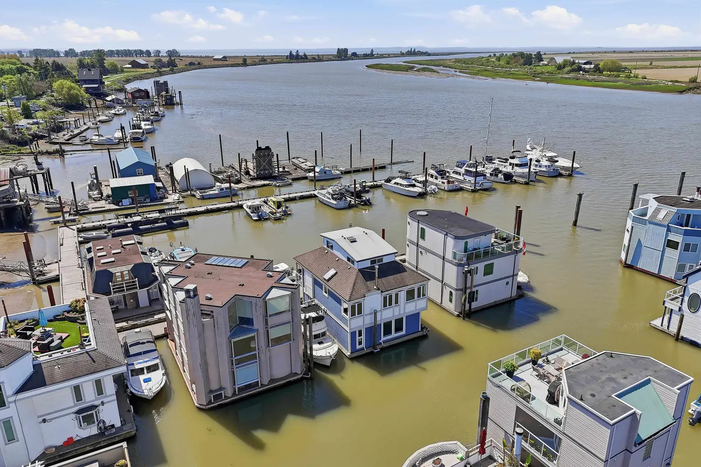 Aerial view of a houseboat community on a wide river, with boats docked nearby. Houses are various colors and styles.
