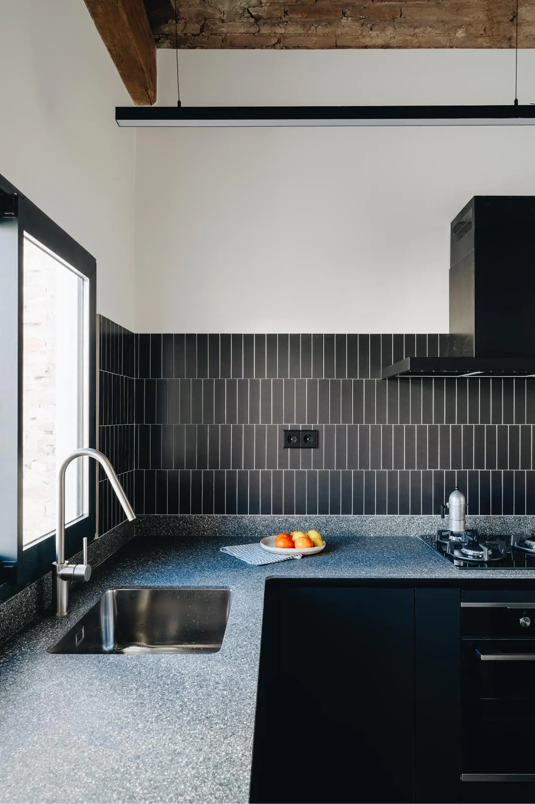 Modern kitchen with black cabinets, speckled countertop, and vertical black tile backsplash. A stainless steel sink and faucet are visible near a window.
