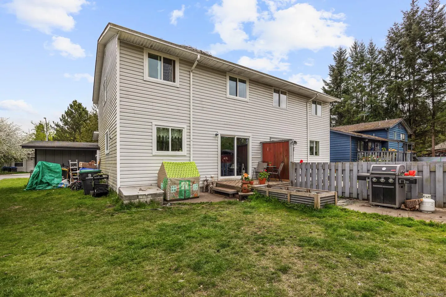 Backyard of a two-story house with light siding, a small deck, a grill, and a wooden fence.