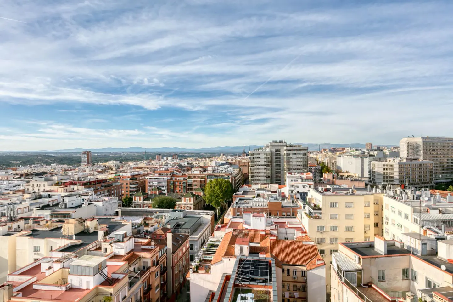 Cityscape view of Madrid, Spain, under a blue sky with wispy clouds. Buildings with red tile roofs and modern high-rises fill the scene.