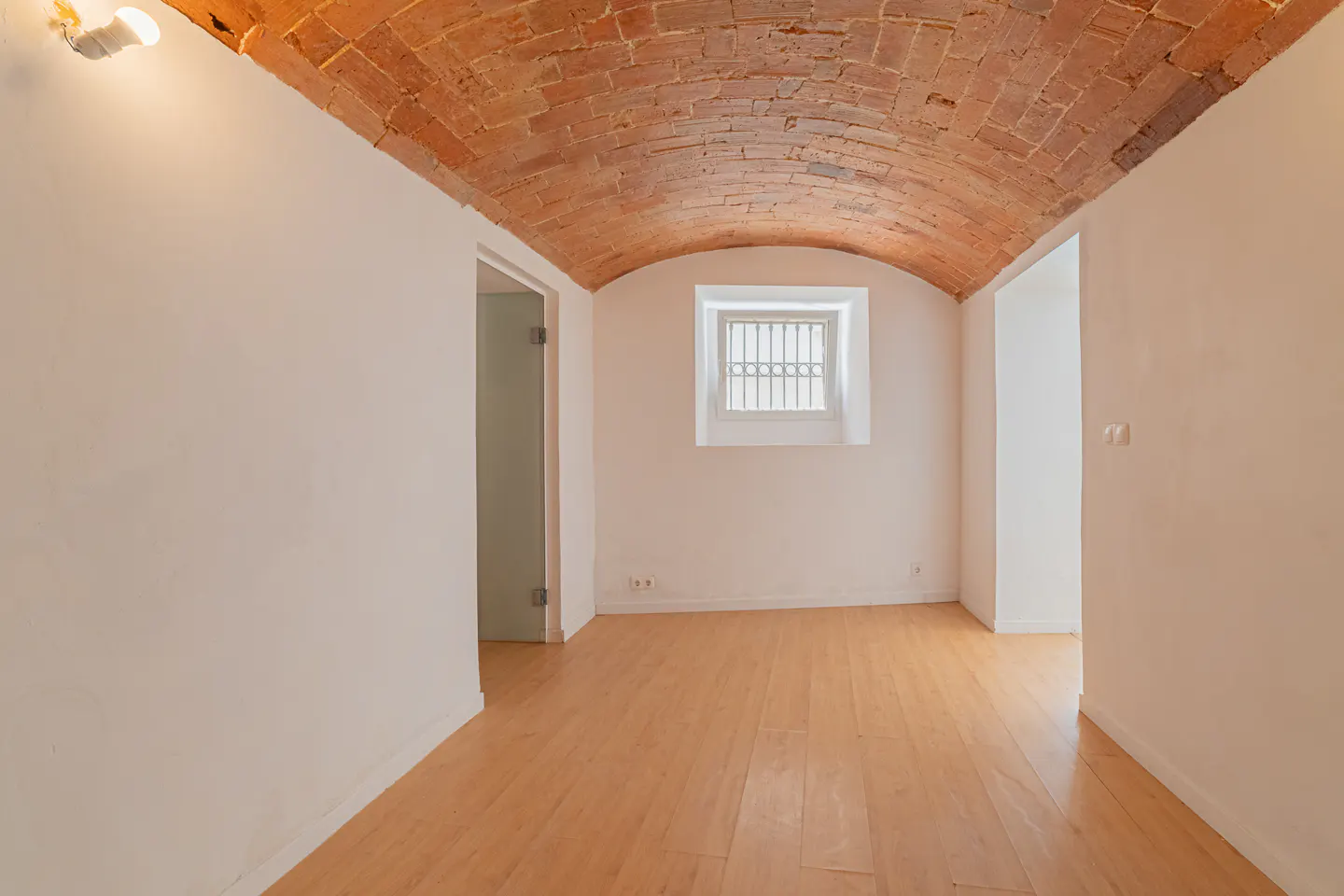 Empty room with a brick vaulted ceiling, light wood floors, white walls, and a barred window.