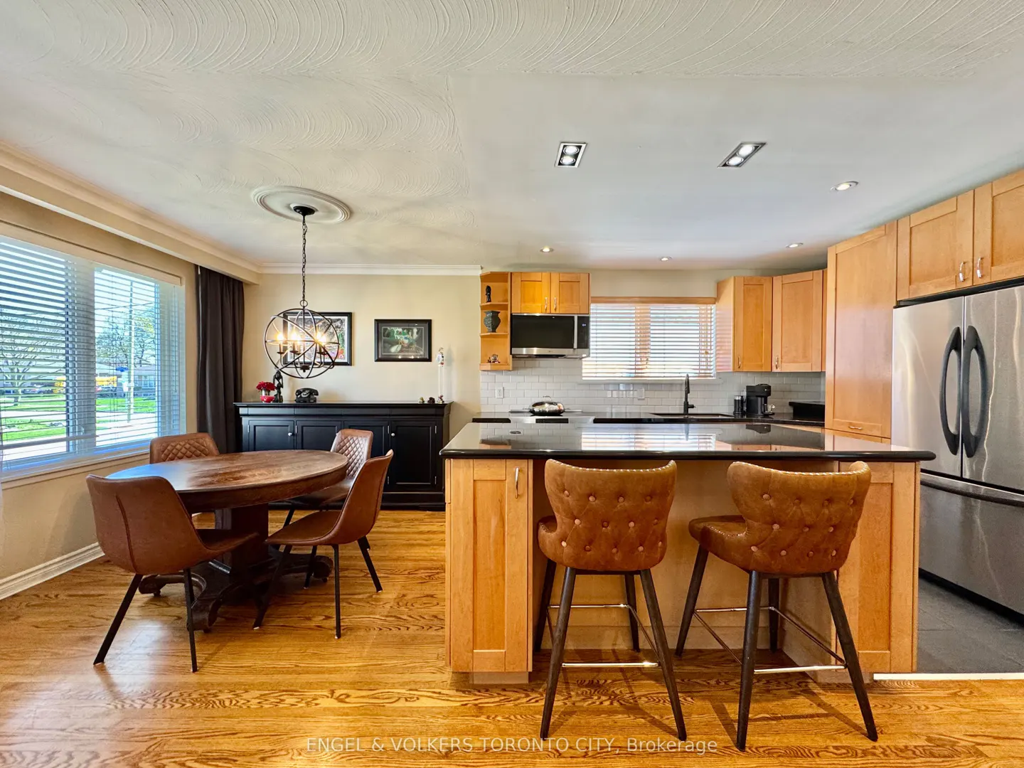 A bright kitchen with wood floors, cabinets, and a black countertop island with two brown leather bar stools. A dining table with four chairs is to the left.