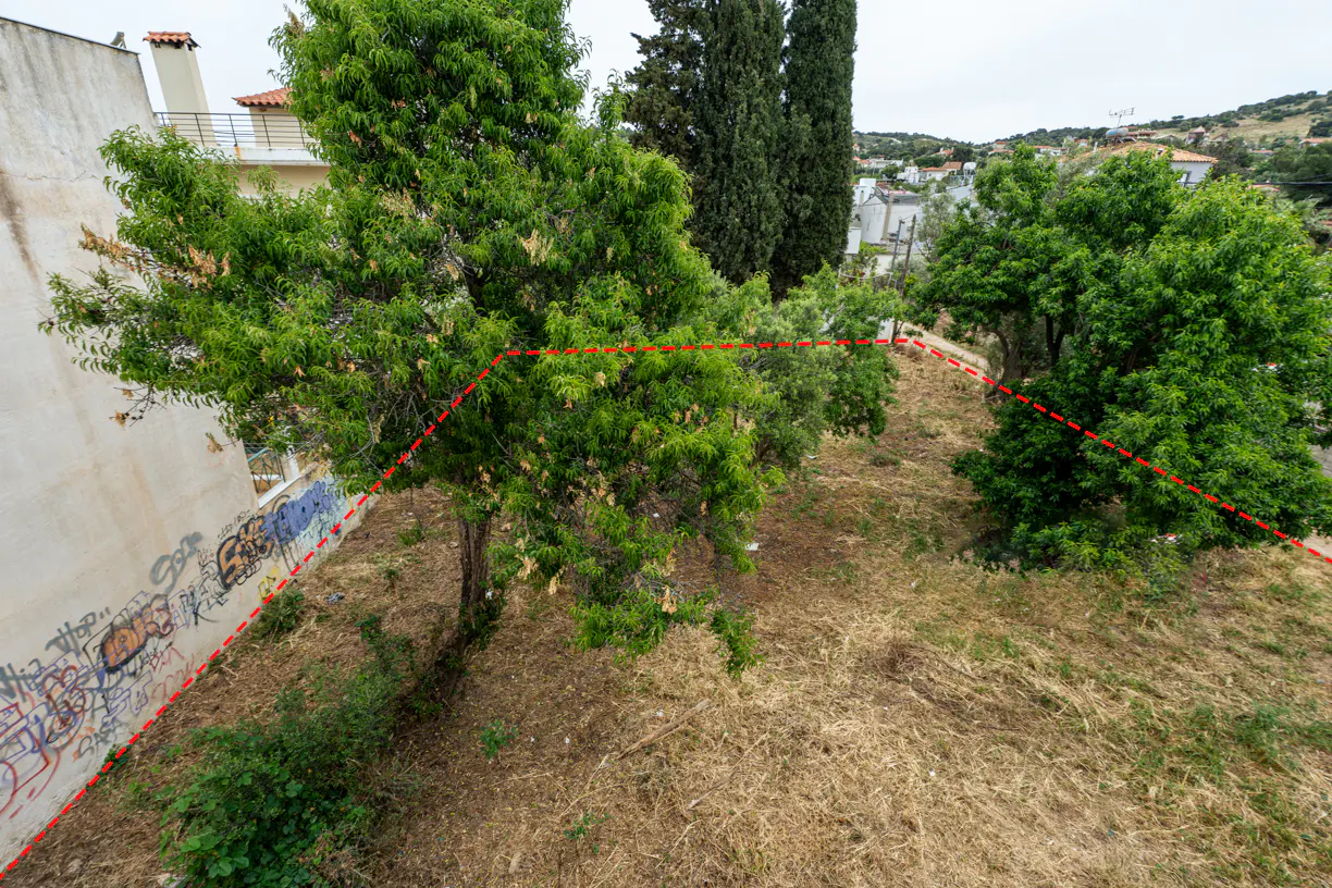 Overhead shot of a vacant lot with trees, dry grass, and a graffiti-covered wall, marked by a red dotted line.