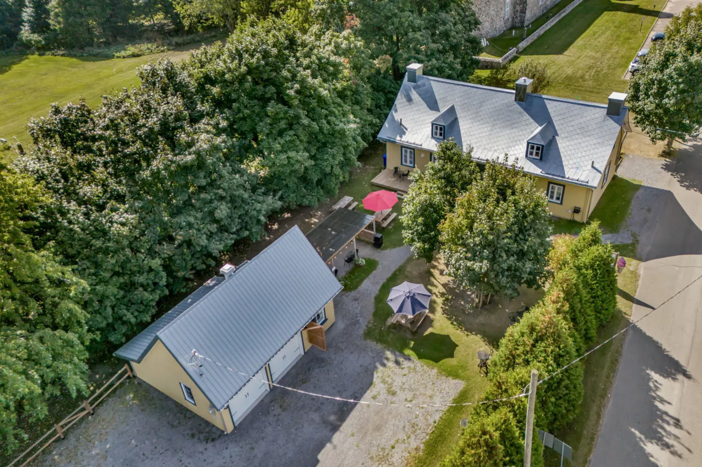 Aerial view of a yellow house with a gray roof, surrounded by trees and a green lawn. A detached garage and outdoor seating area are visible.