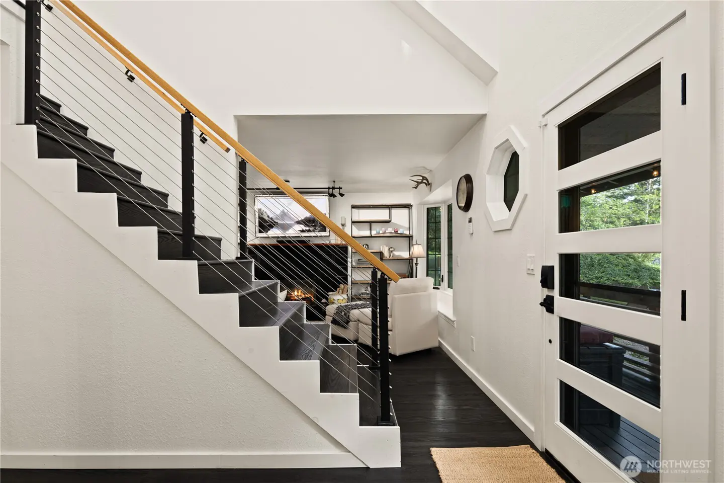 Modern entryway with dark wood stairs, cable railing, and a white front door leading to a cozy living room with fireplace.