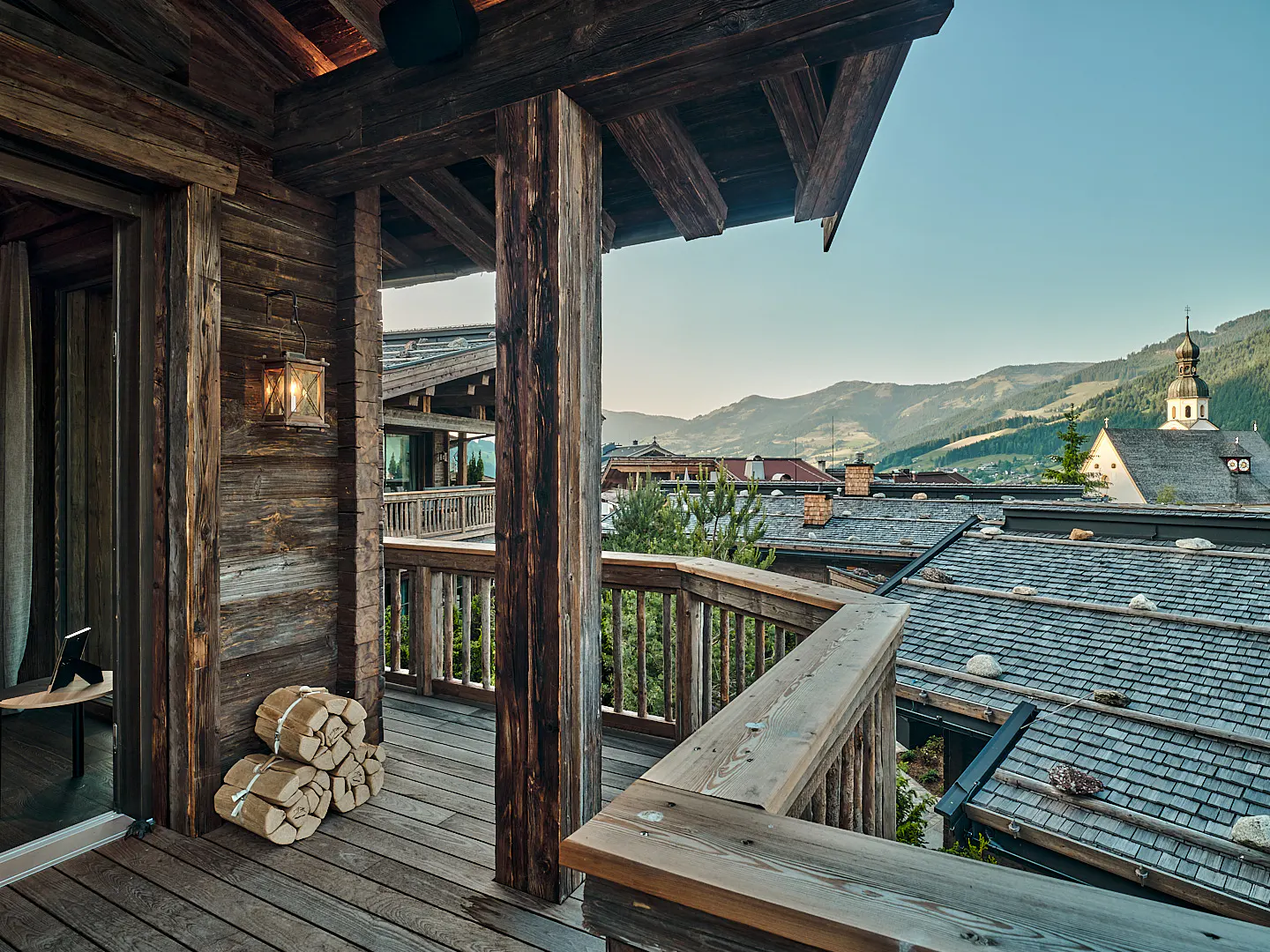 Rustic wood balcony with mountain view. Firewood stacked near an open door. A church steeple rises above tiled roofs.