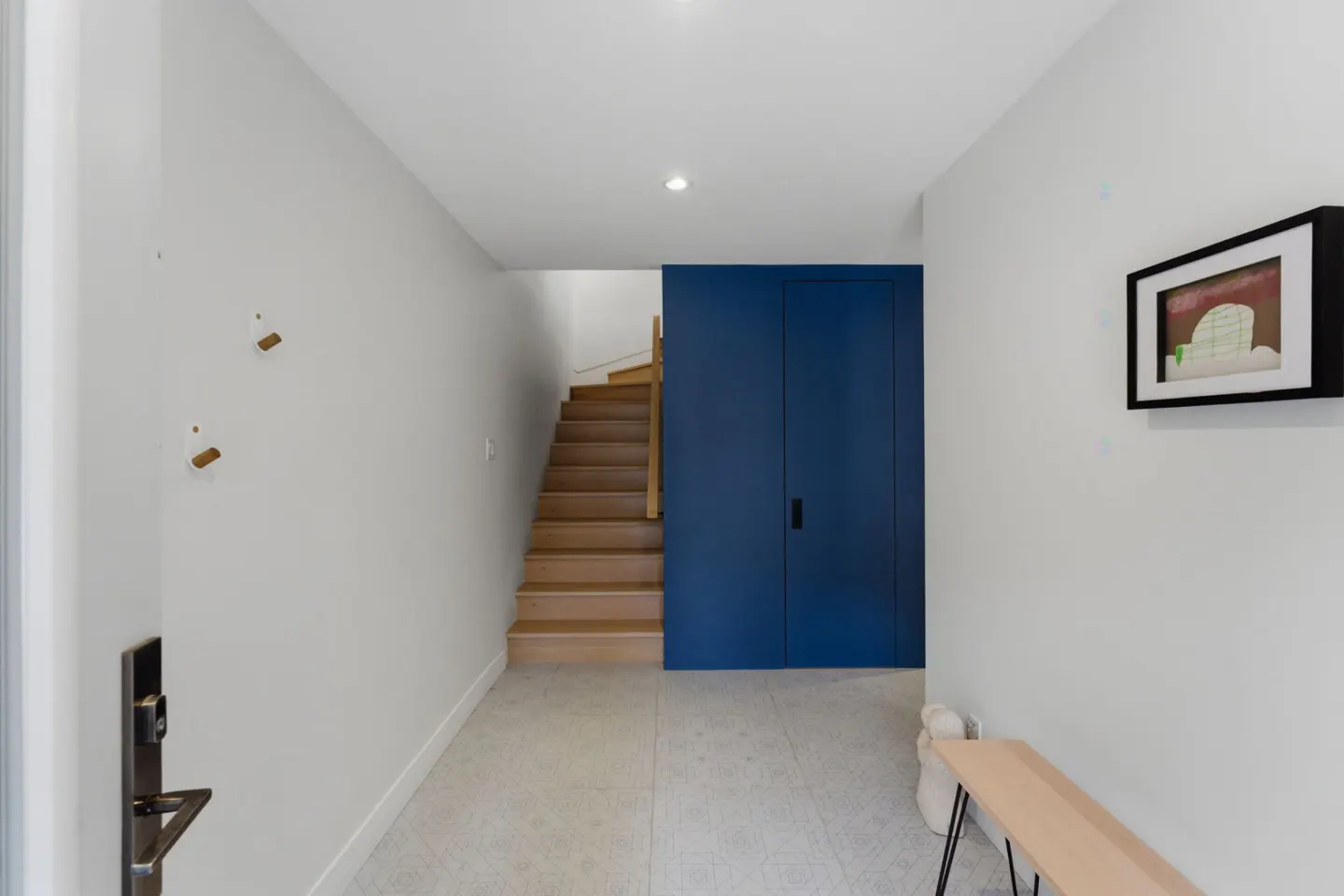 Bright foyer with white walls, patterned tile floor, and wooden stairs. A blue closet door is centered, and a framed picture hangs on the right wall.