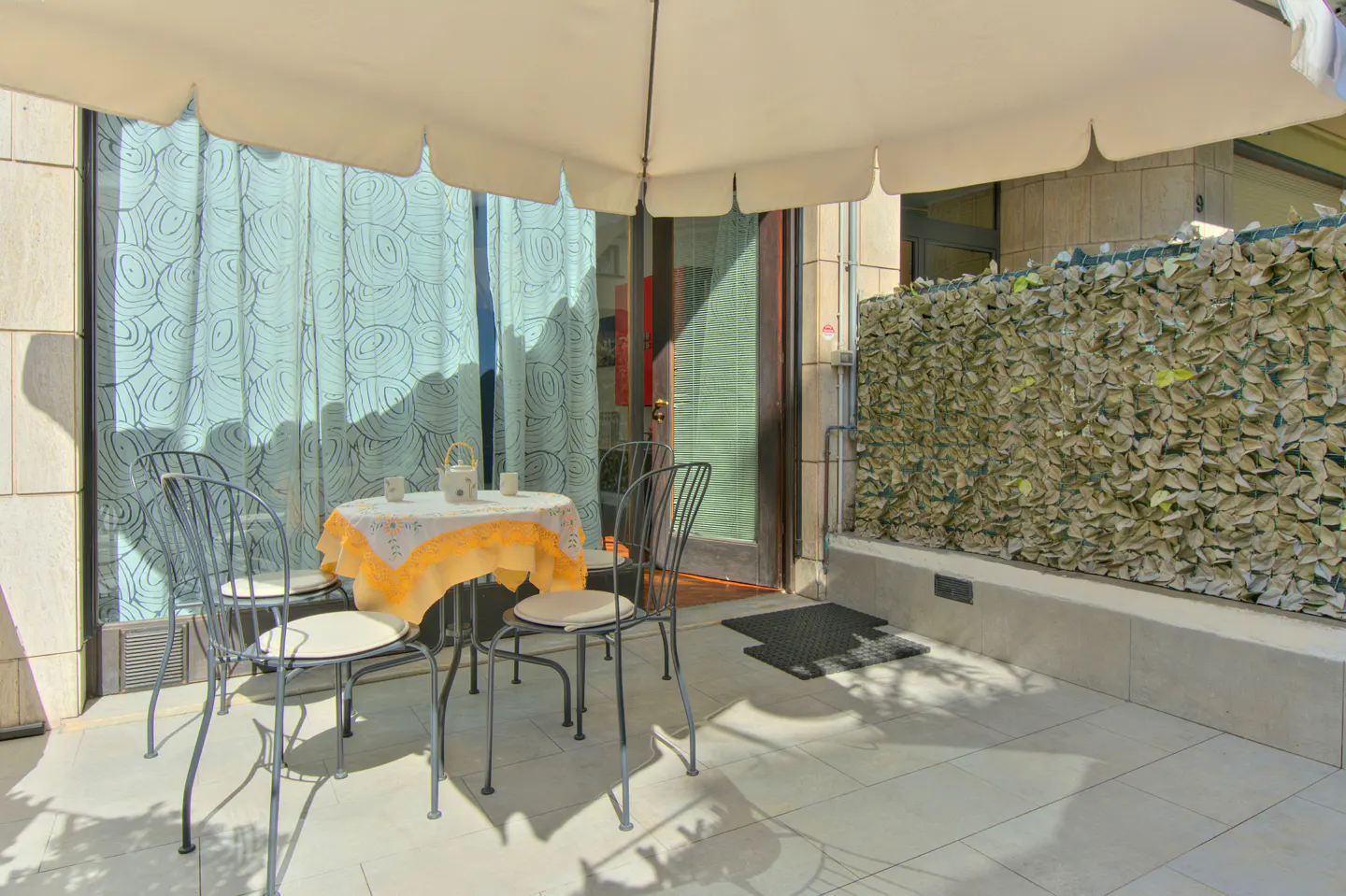Outdoor patio with a table and four chairs under a large umbrella. A door and leafy privacy screen are in the background.