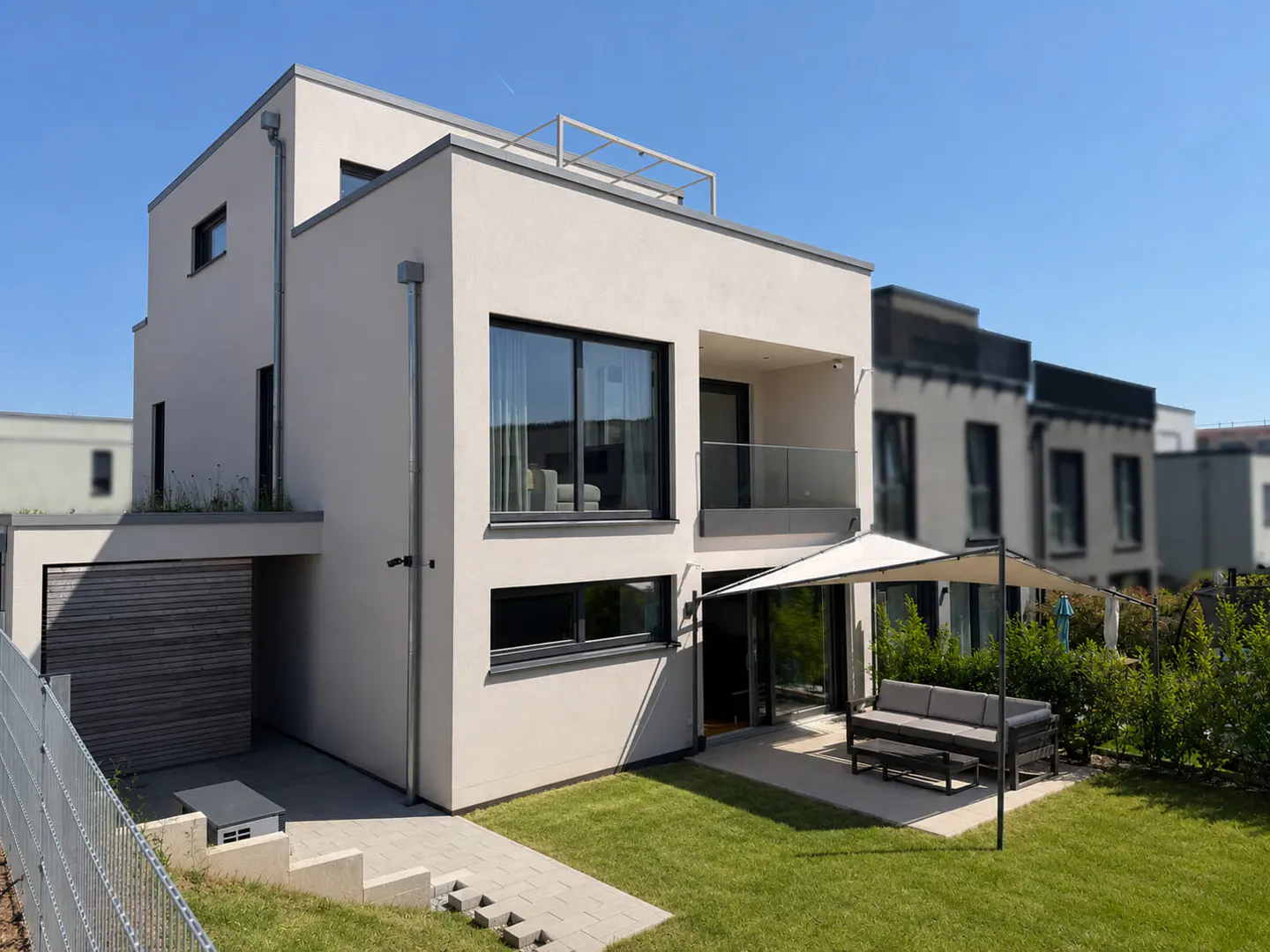 Modern beige two-story house with a flat roof, balcony, patio, and outdoor seating area under a shade sail.