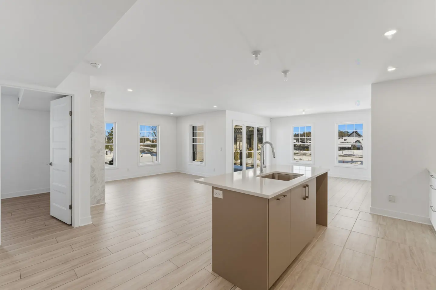 Bright, open-concept kitchen with white walls, light wood floors, and a taupe island with a sink and silver faucet. Windows offer outdoor views.