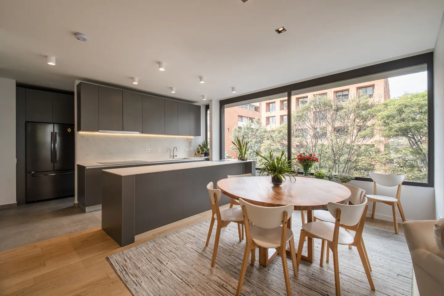 A modern kitchen with gray cabinets, a wood table with white chairs, and a large window with a city view.
